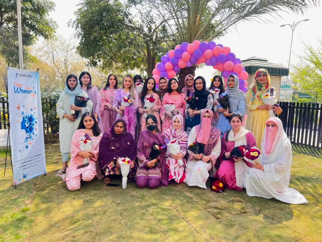 The women of the team holding flowers under a pink and purple balloon arch on Women's Day